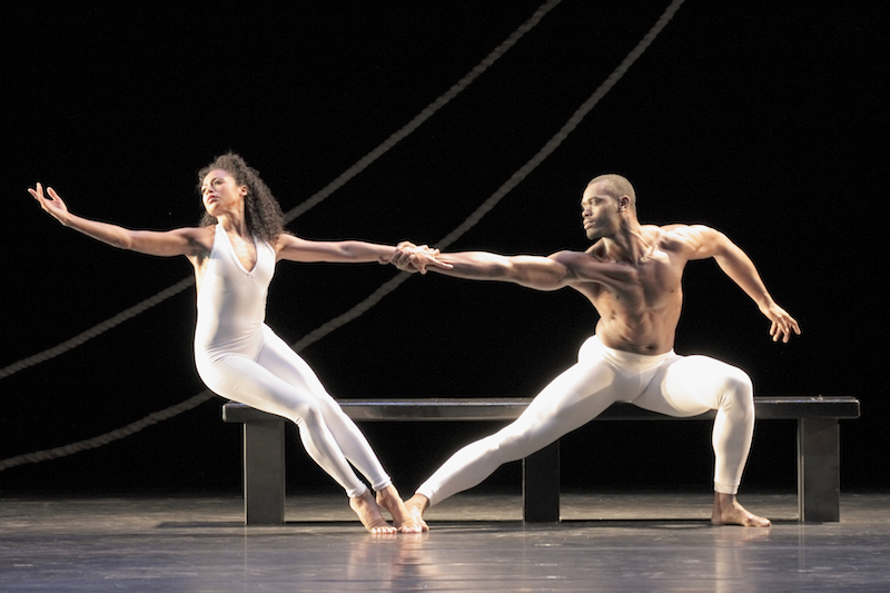 A woman in a white unitard leans precariously to the right while her male parner counter balances her